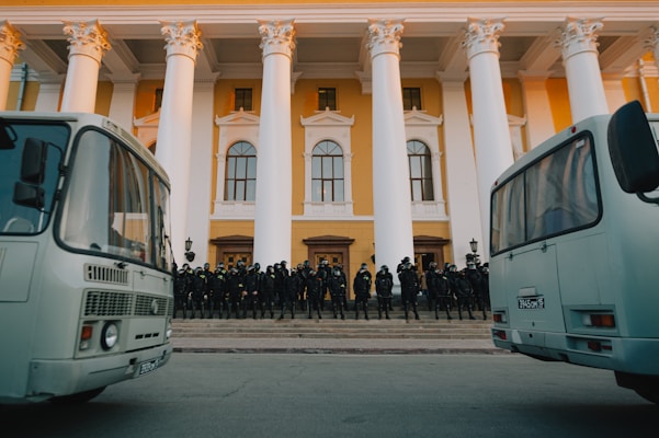A group of uniformed individuals in protective gear, likely police or security personnel, stand in formation on the steps of a large building with classical architecture featuring tall white columns and a yellow facade. Two buses are parked prominently in the foreground.