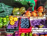 A vibrant display of assorted fruits in a market setting, including green apples, melons, oranges wrapped in netting, pears, and possibly grapefruits or pomelos. The fruits are organized in wooden crates, some with branding. Natural light casts dynamic shadows and highlights the bright colors of the produce.