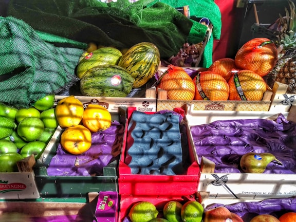 A vibrant display of assorted fruits in a market setting, including green apples, melons, oranges wrapped in netting, pears, and possibly grapefruits or pomelos. The fruits are organized in wooden crates, some with branding. Natural light casts dynamic shadows and highlights the bright colors of the produce.