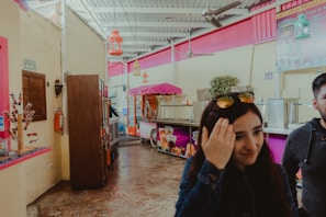 A colorful indoor setting with a vividly decorated ice cream shop. Bright pink and yellow walls with hanging lanterns, refrigerated counters, and a cart with a pink canopy. Two people are present, one in the foreground adjusting sunglasses, and another partially visible in the background.