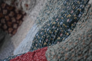A close-up view of a quilt with various floral patterns. The dominant green fabric features small flowers in yellow and white, with hints of red and blue. The quilt appears textured and soft, indicating the presence of stitching and potentially hand-made craftsmanship.
