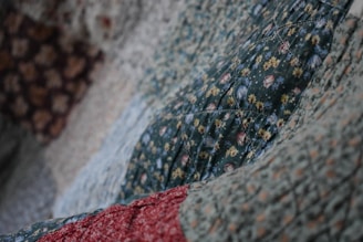 A close-up view of a quilt with various floral patterns. The dominant green fabric features small flowers in yellow and white, with hints of red and blue. The quilt appears textured and soft, indicating the presence of stitching and potentially hand-made craftsmanship.