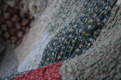 A close-up view of a quilt with various floral patterns. The dominant green fabric features small flowers in yellow and white, with hints of red and blue. The quilt appears textured and soft, indicating the presence of stitching and potentially hand-made craftsmanship.