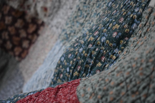 A close-up view of a quilt with various floral patterns. The dominant green fabric features small flowers in yellow and white, with hints of red and blue. The quilt appears textured and soft, indicating the presence of stitching and potentially hand-made craftsmanship.