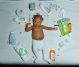 A baby lies on a white quilt surrounded by various baby care products, such as lotions, baby oil, and diapers. The baby is wearing a white diaper with colorful hair ties scattered around. Bright packaging and diverse product shapes create an organized composition.