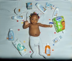 A baby lies on a white quilt surrounded by various baby care products, such as lotions, baby oil, and diapers. The baby is wearing a white diaper with colorful hair ties scattered around. Bright packaging and diverse product shapes create an organized composition.