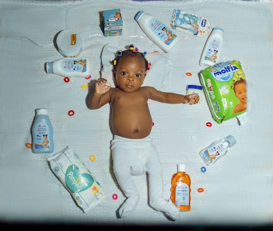 A baby lies on a white quilt surrounded by various baby care products, such as lotions, baby oil, and diapers. The baby is wearing a white diaper with colorful hair ties scattered around. Bright packaging and diverse product shapes create an organized composition.