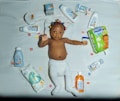 A baby lies on a white quilt surrounded by various baby care products, such as lotions, baby oil, and diapers. The baby is wearing a white diaper with colorful hair ties scattered around. Bright packaging and diverse product shapes create an organized composition.