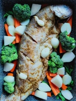 A seasoned whole fish is laid out on a baking tray surrounded by chunks of fresh vegetables, including broccoli, carrots, and onions. The fish appears to be marinated, showing a light brown seasoning on its skin. The vegetables are neatly arranged around the fish, adding a vibrant touch to the overall composition.