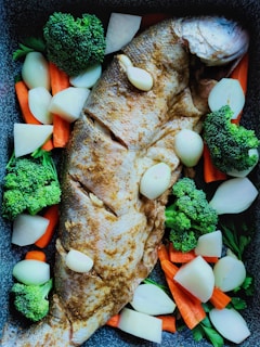 A seasoned whole fish is laid out on a baking tray surrounded by chunks of fresh vegetables, including broccoli, carrots, and onions. The fish appears to be marinated, showing a light brown seasoning on its skin. The vegetables are neatly arranged around the fish, adding a vibrant touch to the overall composition.