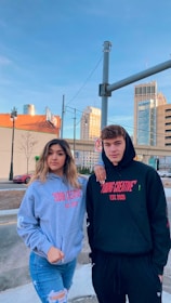 Two young individuals stand outdoors in an urban setting, both wearing hoodies with the text 'YOUNG CREATIVE EST. 2020' printed on them. The background showcases buildings, a street, and a clear blue sky. A woman wears a gray hoodie and jeans, and a man wears a black hoodie and black pants.