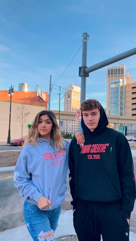 Two young individuals stand outdoors in an urban setting, both wearing hoodies with the text 'YOUNG CREATIVE EST. 2020' printed on them. The background showcases buildings, a street, and a clear blue sky. A woman wears a gray hoodie and jeans, and a man wears a black hoodie and black pants.