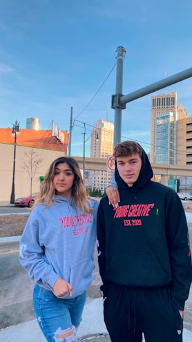 Two young individuals stand outdoors in an urban setting, both wearing hoodies with the text 'YOUNG CREATIVE EST. 2020' printed on them. The background showcases buildings, a street, and a clear blue sky. A woman wears a gray hoodie and jeans, and a man wears a black hoodie and black pants.