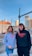 Two young individuals stand outdoors in an urban setting, both wearing hoodies with the text 'YOUNG CREATIVE EST. 2020' printed on them. The background showcases buildings, a street, and a clear blue sky. A woman wears a gray hoodie and jeans, and a man wears a black hoodie and black pants.