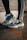 A pair of blue and white athletic shoes are worn by someone standing on a wet concrete surface. The person is wearing black leggings with white stripes on the side. A slight reflection is visible on the ground due to the water.