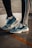 A pair of blue and white athletic shoes are worn by someone standing on a wet concrete surface. The person is wearing black leggings with white stripes on the side. A slight reflection is visible on the ground due to the water.