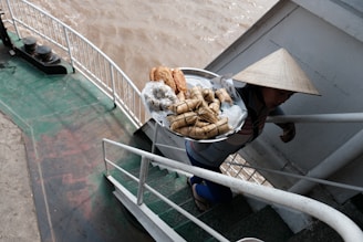 A person wearing a traditional conical hat is ascending a set of stairs on a boat, carrying a tray filled with a variety of wrapped foods and pastries. The background shows part of the boat and a view of the brown, choppy river water.