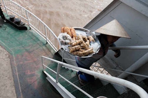 A person wearing a traditional conical hat is ascending a set of stairs on a boat, carrying a tray filled with a variety of wrapped foods and pastries. The background shows part of the boat and a view of the brown, choppy river water.