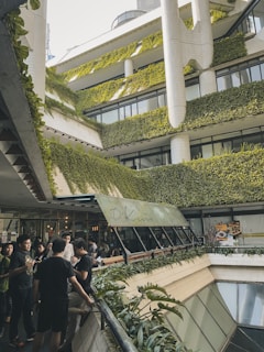 A multi-level building adorned with lush greenery on its exterior, creating a vertical garden effect. People gather in an open area, with a restaurant sign visible, indicating a social setting. Architectural design is modern with clean lines and extensive plant coverage.