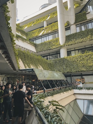 A multi-level building adorned with lush greenery on its exterior, creating a vertical garden effect. People gather in an open area, with a restaurant sign visible, indicating a social setting. Architectural design is modern with clean lines and extensive plant coverage.