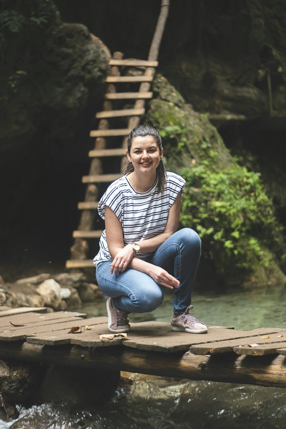 woman in white and black stripe shirt and blue denim jeans sitting on brown wooden bridge