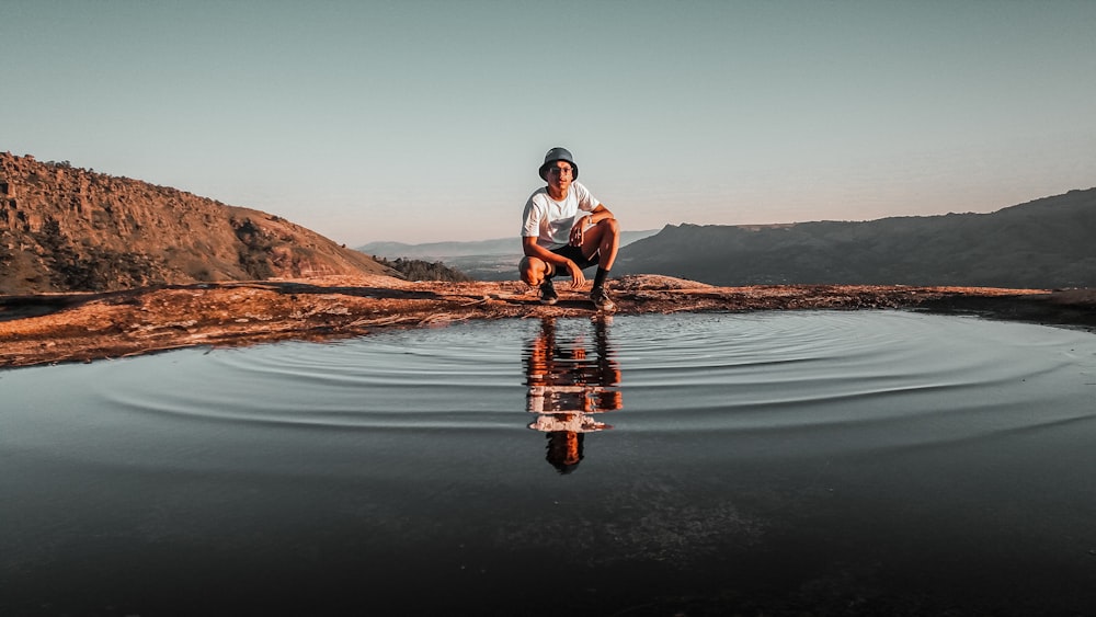 man in white t-shirt and blue denim shorts sitting on rock in front of lake