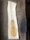 A slice of light-colored wood sits atop a darker wooden surface. The slice shows a natural grain pattern, and there is an irregular-shaped stain or mark in a slightly darker shade of brown on it. The surface beneath the wood slice has a rough texture.
