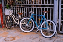 Two bicycles are leaning against a metal fence outdoors. One bicycle is cream-colored with a brown seat and a front basket, while the other is blue with a brown seat. The ground is paved with tiles, and there is some sand scattered around.
