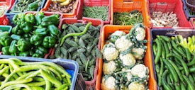 A variety of fresh vegetables displayed in colorful baskets, including bell peppers, bitter gourds, cauliflowers, cucumbers, beans, and ginger. The vibrant and diverse collection is neatly organized.