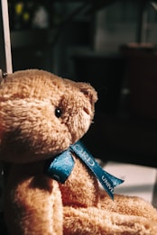 A plush teddy bear with a soft, furry texture is adorned with a blue ribbon tied around its neck, featuring inscribed text. The bear is positioned in a warmly lit environment, with a blurred background that suggests an indoor setting.