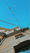 An upward perspective of a building, showcasing architectural details against a clear blue sky. Multiple electrical cables crisscross above, adorned with small, glowing lights. The building's facade features decorative molding and a mix of beige and brown tones.