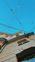 An upward perspective of a building, showcasing architectural details against a clear blue sky. Multiple electrical cables crisscross above, adorned with small, glowing lights. The building's facade features decorative molding and a mix of beige and brown tones.