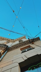 An upward perspective of a building, showcasing architectural details against a clear blue sky. Multiple electrical cables crisscross above, adorned with small, glowing lights. The building's facade features decorative molding and a mix of beige and brown tones.