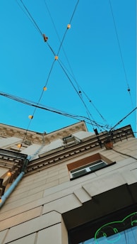 An upward perspective of a building, showcasing architectural details against a clear blue sky. Multiple electrical cables crisscross above, adorned with small, glowing lights. The building's facade features decorative molding and a mix of beige and brown tones.