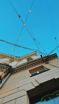 An upward perspective of a building, showcasing architectural details against a clear blue sky. Multiple electrical cables crisscross above, adorned with small, glowing lights. The building's facade features decorative molding and a mix of beige and brown tones.