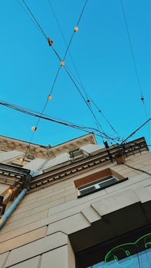 An upward perspective of a building, showcasing architectural details against a clear blue sky. Multiple electrical cables crisscross above, adorned with small, glowing lights. The building's facade features decorative molding and a mix of beige and brown tones.