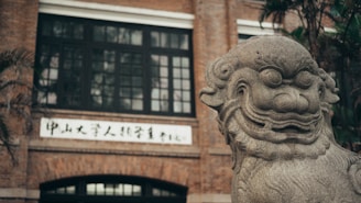A detailed stone statue of a traditional Chinese mythical creature in the foreground, located in front of a large brick building with multiple windows and Chinese characters on a white signboard. The greenery in the background adds a touch of nature.