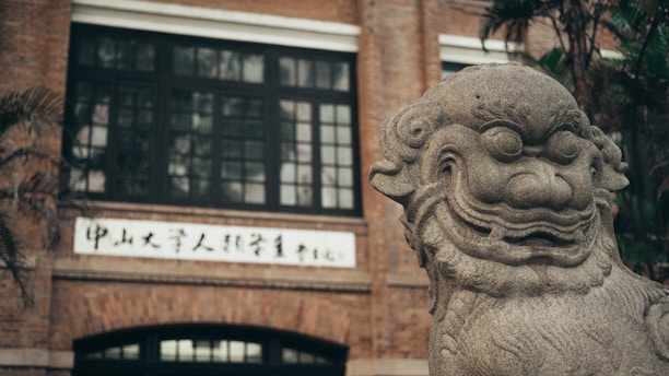A detailed stone statue of a traditional Chinese mythical creature in the foreground, located in front of a large brick building with multiple windows and Chinese characters on a white signboard. The greenery in the background adds a touch of nature.