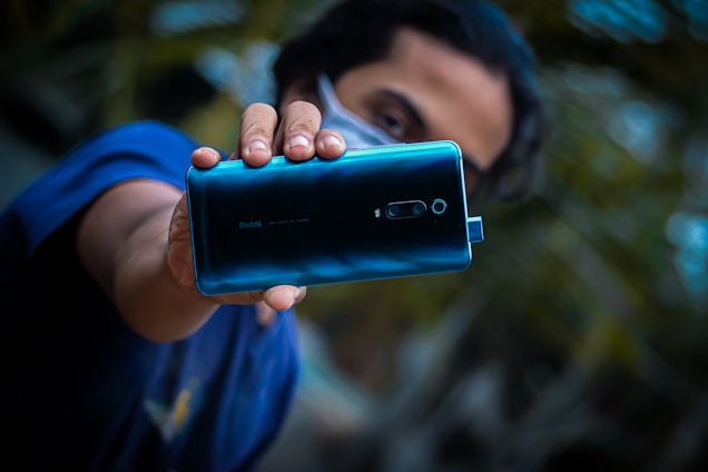 A person holding a blue Redmi smartphone closely to the camera, showing the back of the phone. The background is blurred with some greenery visible. The person is wearing a dark blue shirt and a face mask.