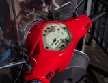 A close-up view of a red scooter's dashboard featuring a speedometer with various colored lights embedded. The handlebars are visible, and the scooter is parked on a pavement with railings in the background.