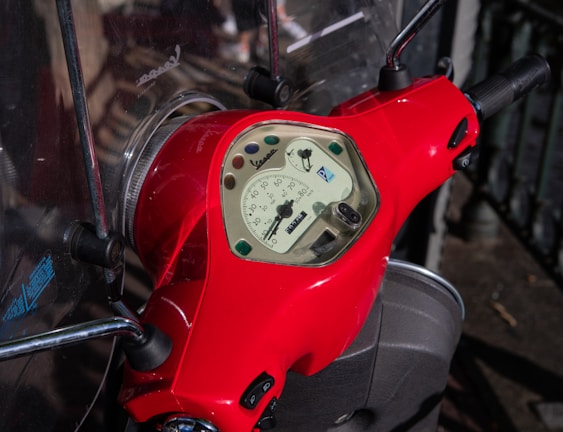 A close-up view of a red scooter's dashboard featuring a speedometer with various colored lights embedded. The handlebars are visible, and the scooter is parked on a pavement with railings in the background.