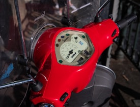 A close-up view of a red scooter's dashboard featuring a speedometer with various colored lights embedded. The handlebars are visible, and the scooter is parked on a pavement with railings in the background.