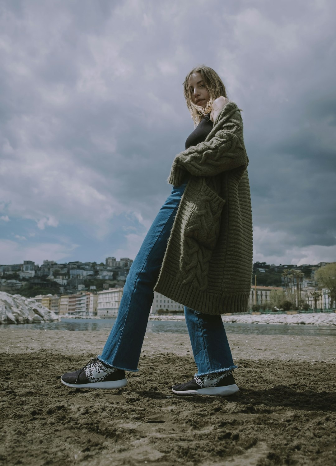 woman in gray coat and blue denim jeans standing on gray sand during daytime