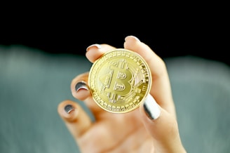 A hand holding a gold-colored coin with the Bitcoin logo prominently displayed on it. The nails are painted in metallic shades, and the background is blurred.