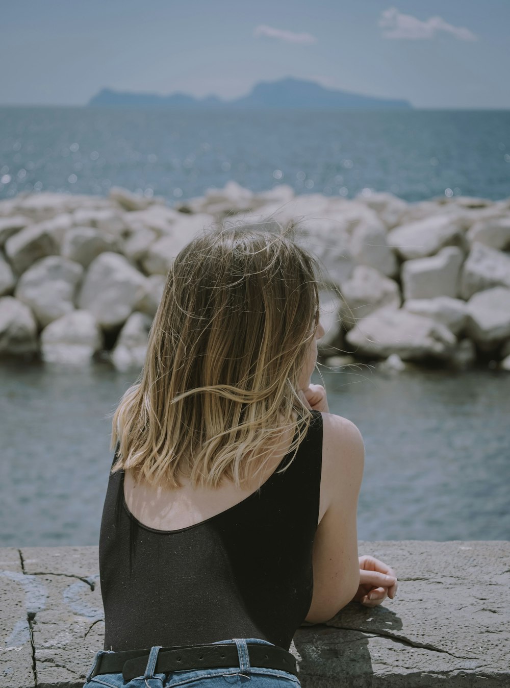 woman in black tank top sitting on gray concrete pavement near body of water during daytime