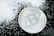 A silver-colored Bitcoin coin lies on a dark surface scattered with small white granules, creating a contrast between the metallic shine of the coin and the matte texture of the granules.