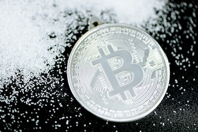 A silver-colored Bitcoin coin lies on a dark surface scattered with small white granules, creating a contrast between the metallic shine of the coin and the matte texture of the granules.