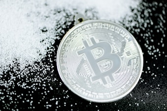 A silver-colored Bitcoin coin lies on a dark surface scattered with small white granules, creating a contrast between the metallic shine of the coin and the matte texture of the granules.