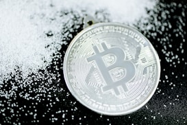 A silver-colored Bitcoin coin lies on a dark surface scattered with small white granules, creating a contrast between the metallic shine of the coin and the matte texture of the granules.