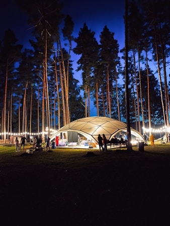 A nighttime setting in a forest with tall pine trees surrounding a well-lit dome-shaped tent. Several people are gathered around, creating a lively atmosphere under strings of lights. The deep blue sky adds a peaceful contrast to the warm yellow lights of the tent.